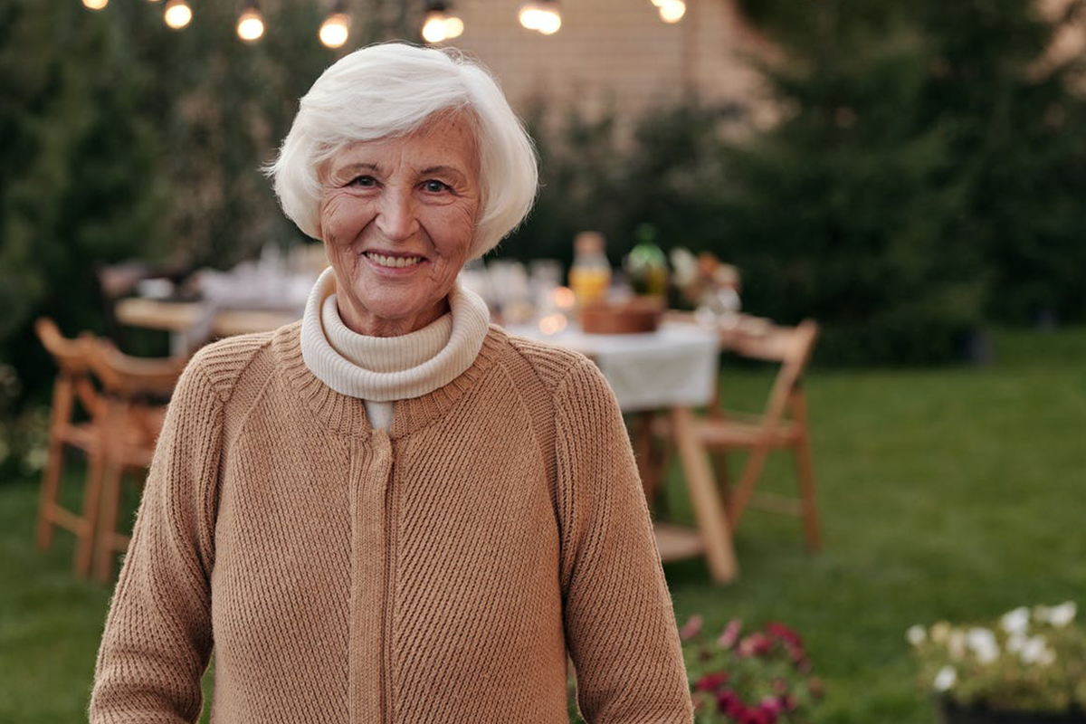 Smiling older woman standing outdoors, showing bright eyes and a refreshed look that can result from eyelid surgery.