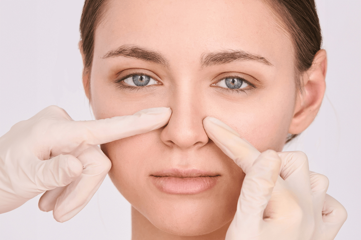 Young woman’s nose being examined by gloved hands, illustrating evaluation for rhinoplasty candidacy and facial balance.