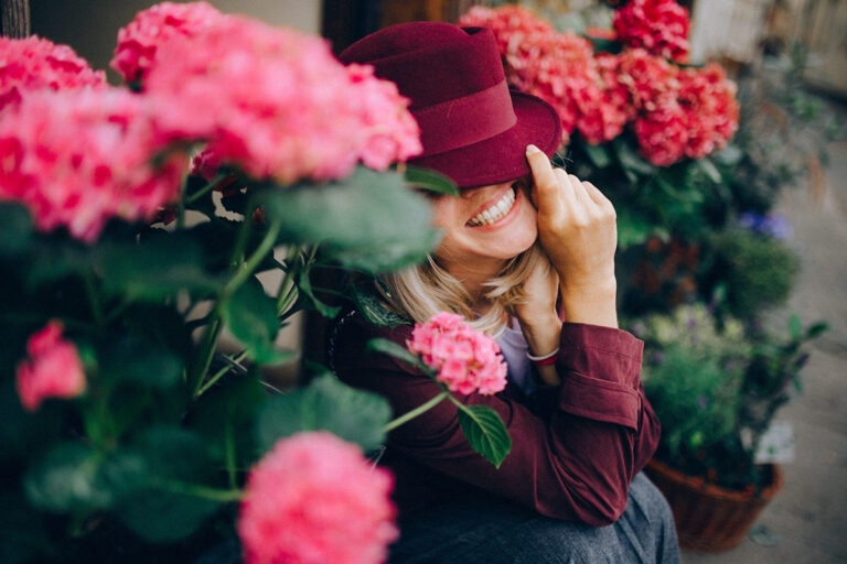Smiling woman in burgundy hat surrounded by pink flowers, symbolizing confidence and comfort following labiaplasty recovery.