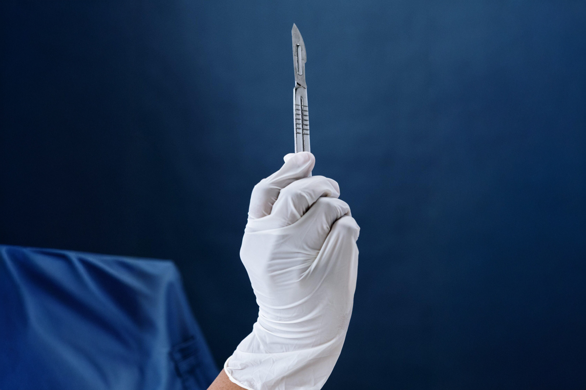 Surgeon’s gloved hand holding a scalpel against a dark blue background, symbolizing cosmetic surgery safety.