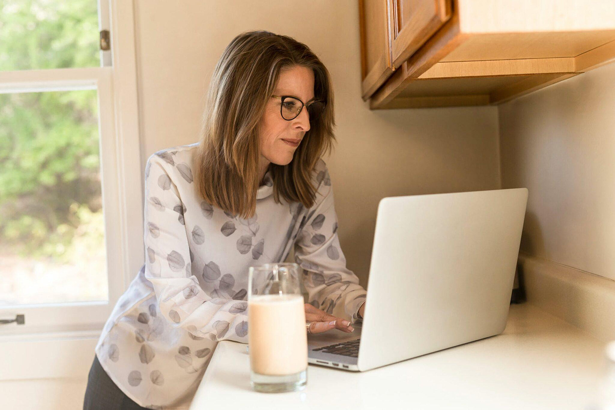 Woman wearing glasses using a laptop in a bright kitchen, representing someone researching brow lift surgery recovery information