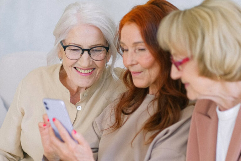 Three older women smiling and looking at a smartphone together, representing shared awareness and discussion of cataract symptoms and eye health