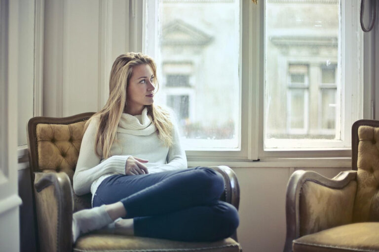 Woman sitting thoughtfully by window in cozy chair, representing preparation and reflection before cosmetic surgery consultation