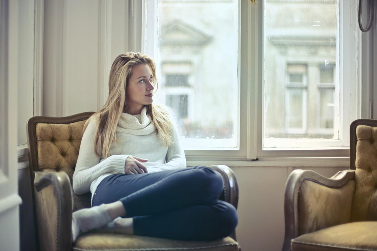 Woman sitting thoughtfully by window in cozy chair, representing preparation and reflection before cosmetic surgery consultation