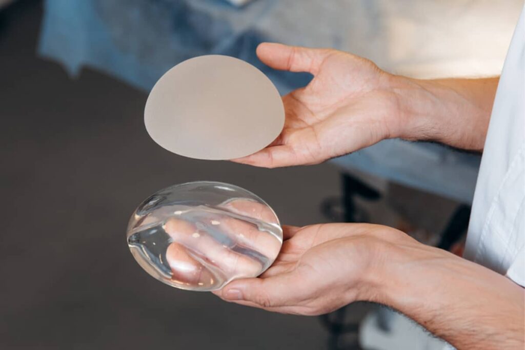 A close-up of a person's hands holding two different breast implants: a textured, frosted silicone gel implant and a smooth, clear saline implant.