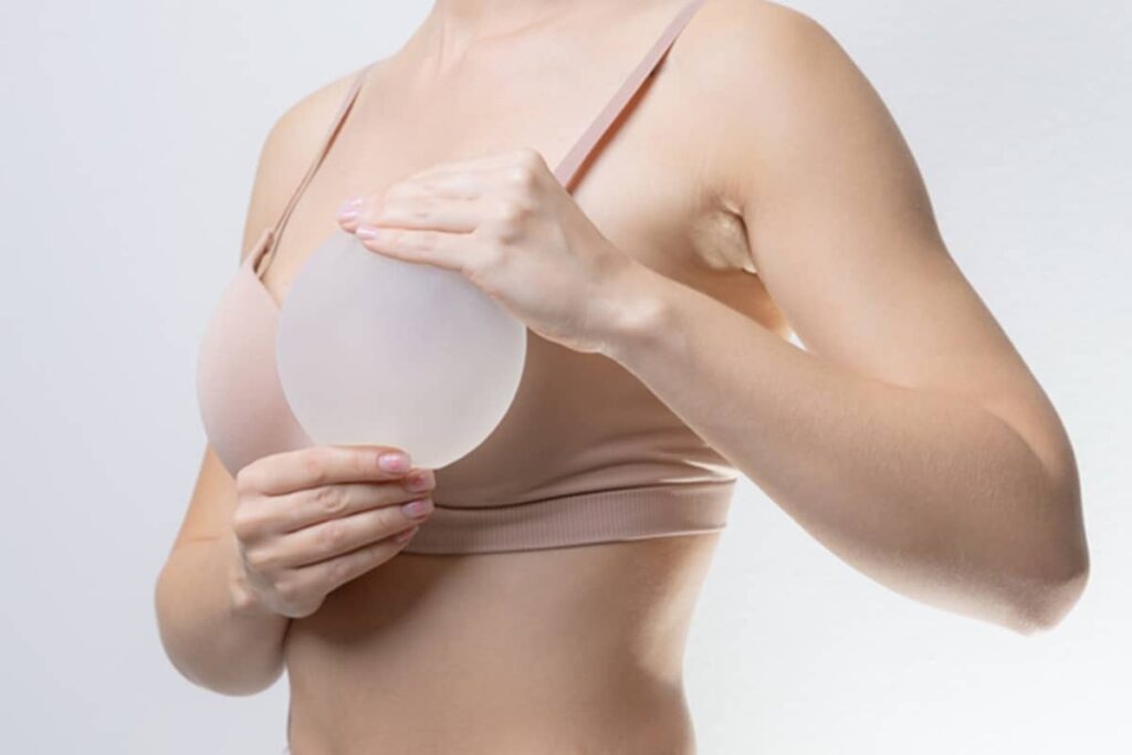 A woman in a tan sports bra holding a round, frosted silicone breast implant in front of her chest to visualize the size.