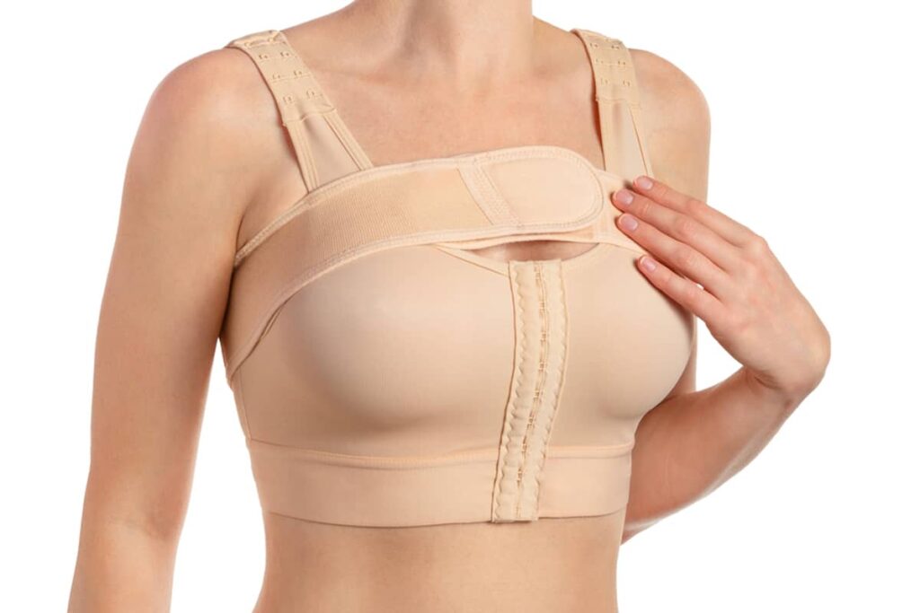 A woman wearing a specialized tan surgical compression bra designed to provide support and minimize tension on healing incisions after breast augmentation.