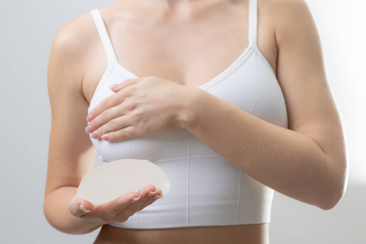 A woman in a white sports bra holding a silicone breast implant, illustrating the relationship between implant size and surgical incision length.