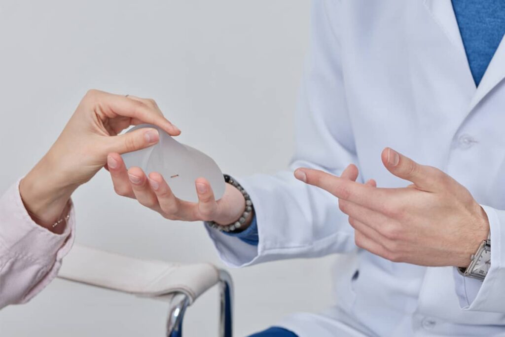 A surgeon in a white lab coat pointing to a silicone implant held by a patient during a consultation.