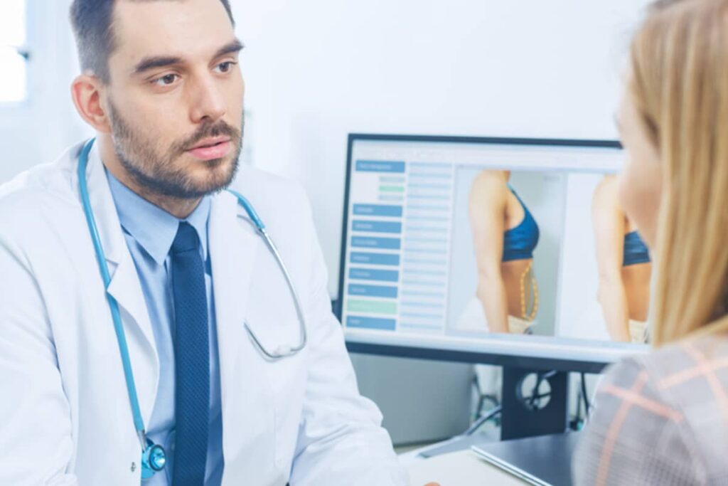 A male doctor in a white coat with a stethoscope discusses body contouring results with a patient, with a computer monitor showing patient profile comparisons in the background.