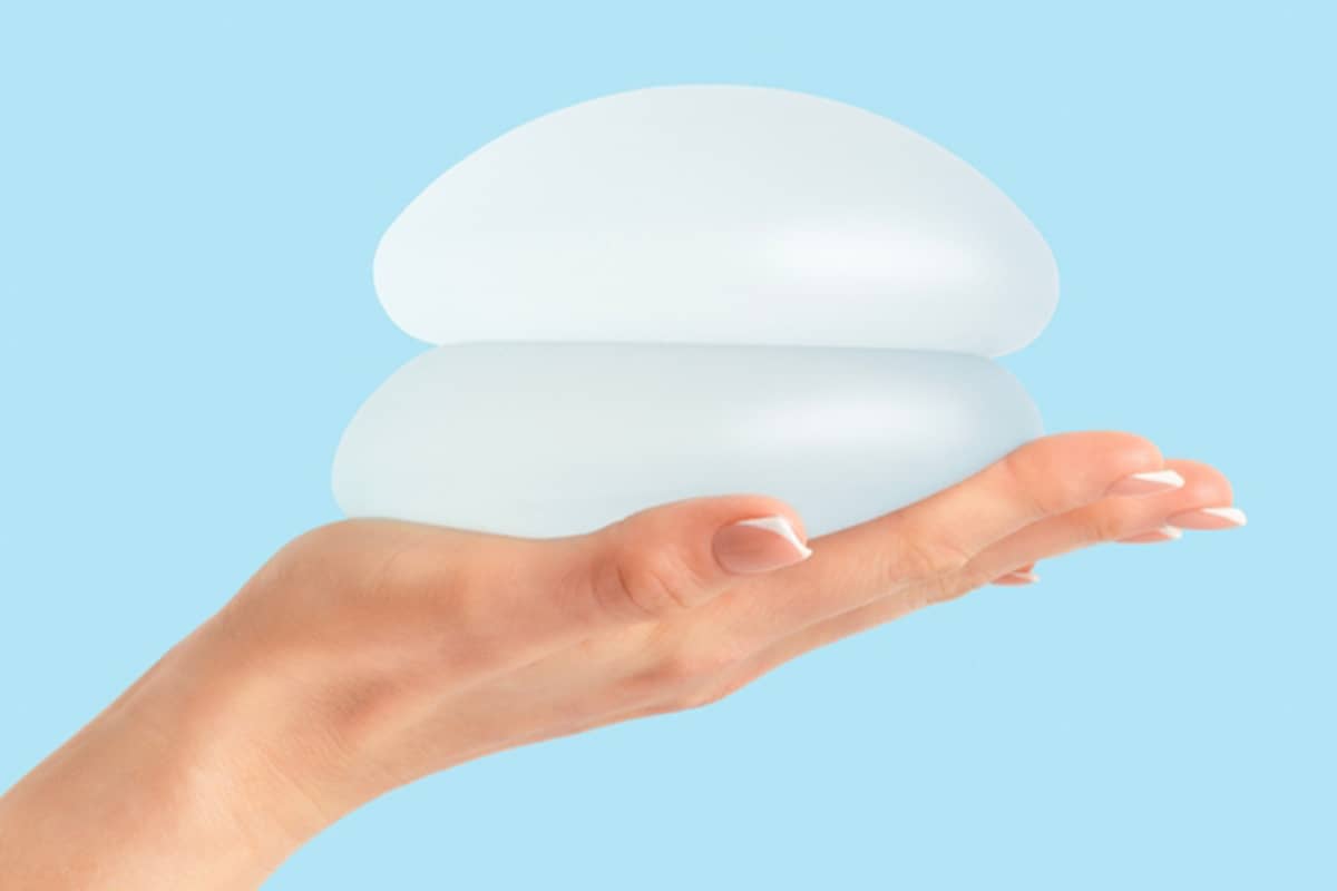 A side profile of a hand holding two stacked, round silicone breast implants against a solid light blue background.