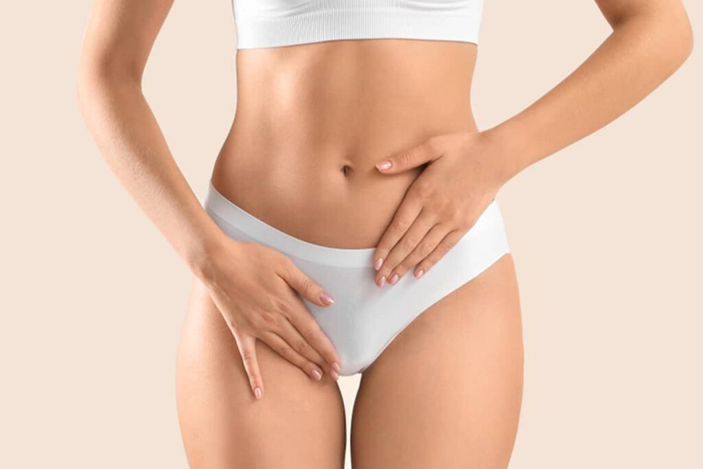 A woman in white lingerie placing her hands over her lower abdomen to evaluate her comfort and physical confidence following a labiaplasty procedure.