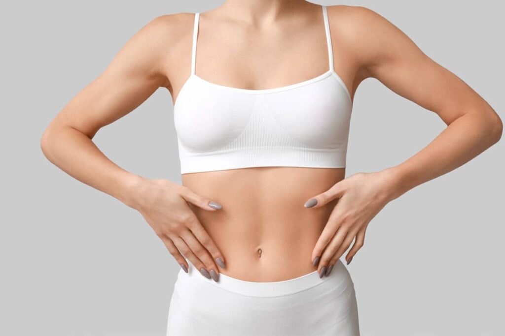 A woman in a white sports bra gently placing her hands on her waist to evaluate her skin texture and healing progress after surgery.