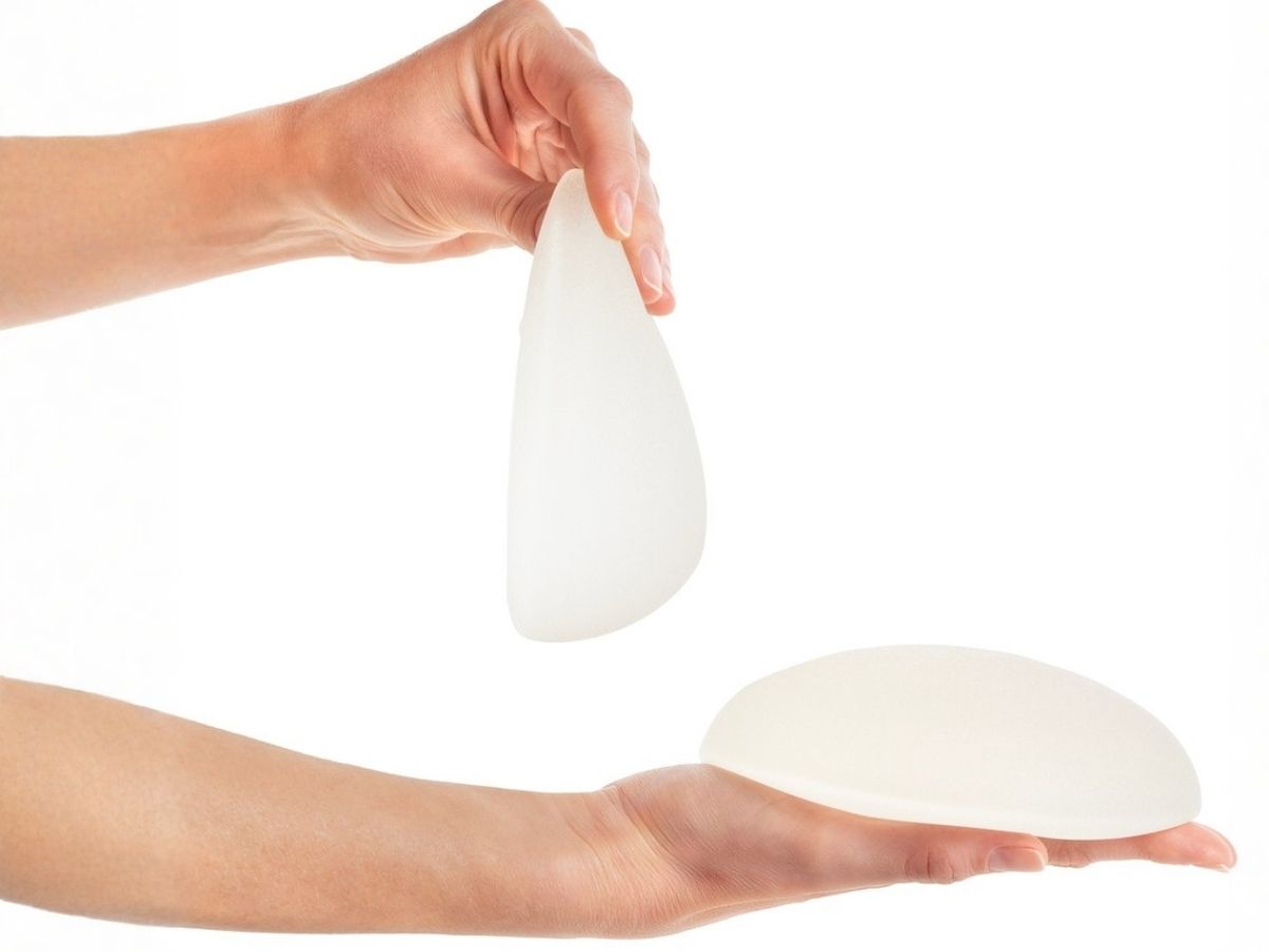 A person’s hands holding a teardrop-shaped silicone implant vertically to show its tapered profile alongside a round implant resting flat.