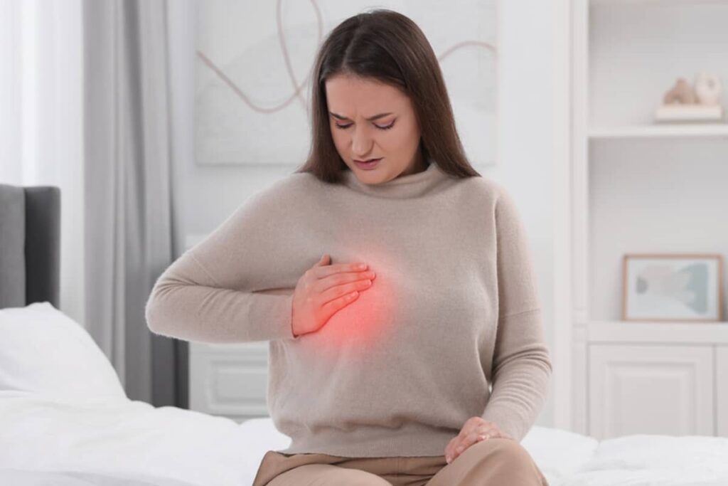 A woman sitting on a bed with a pained expression, holding her chest where a red glow indicates localized pain.