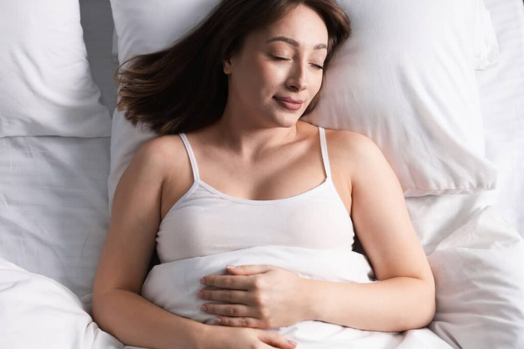 An overhead view of a woman sleeping comfortably on her back in bed with white linens, resting her hands on her stomach.