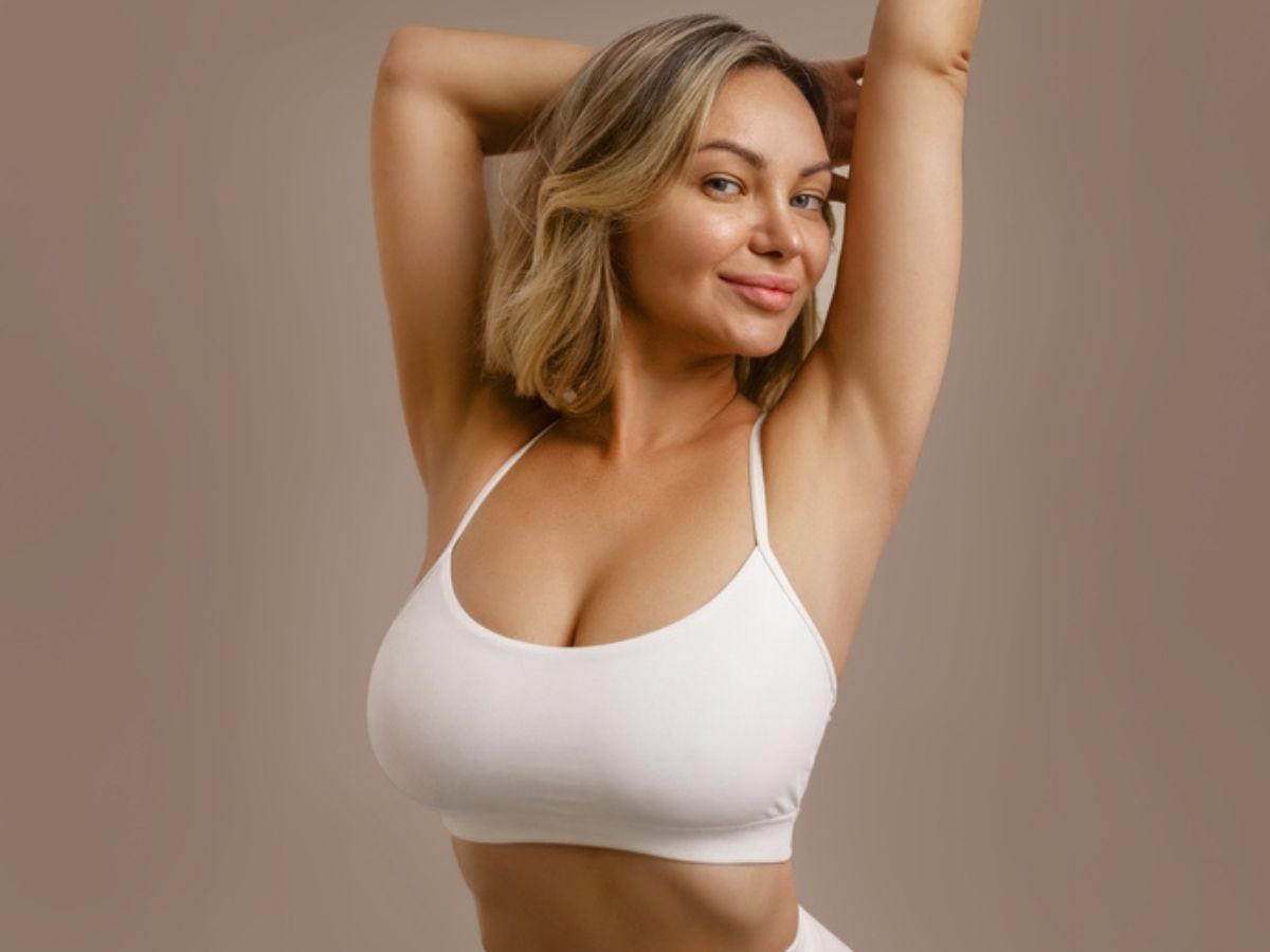 A woman with blonde hair and a white sports bra smiling while posing with her arms raised against a neutral background.