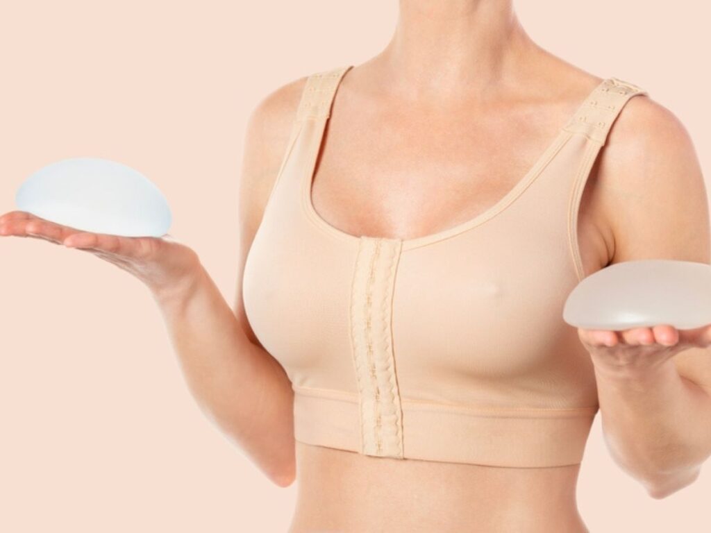 A woman in a tan surgical bra holding a round silicone breast implant in each hand to compare different sizes.