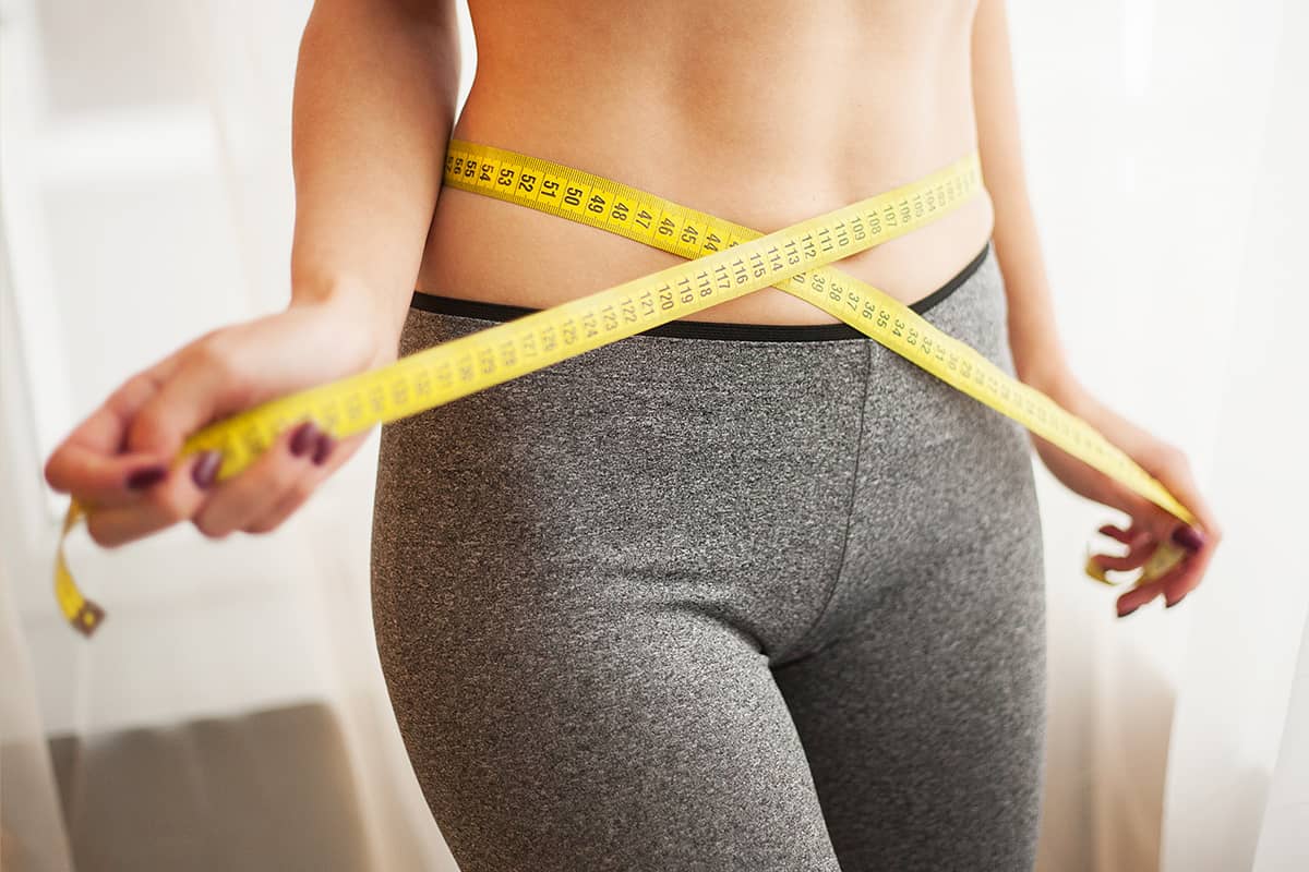 Close-up of a woman in grey leggings measuring her slender waist with a yellow tape measure.