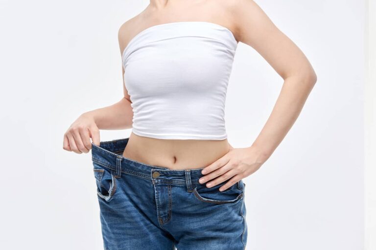 Close-up of a woman's slender torso wearing a white bandeau top, pulling the front of very large, baggy jeans to demonstrate substantial volume loss around her waist.