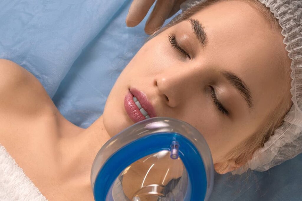 An overhead view of a woman with closed eyes wearing a surgical cap, lying down with a clear and blue anesthesia oxygen mask positioned near her face.