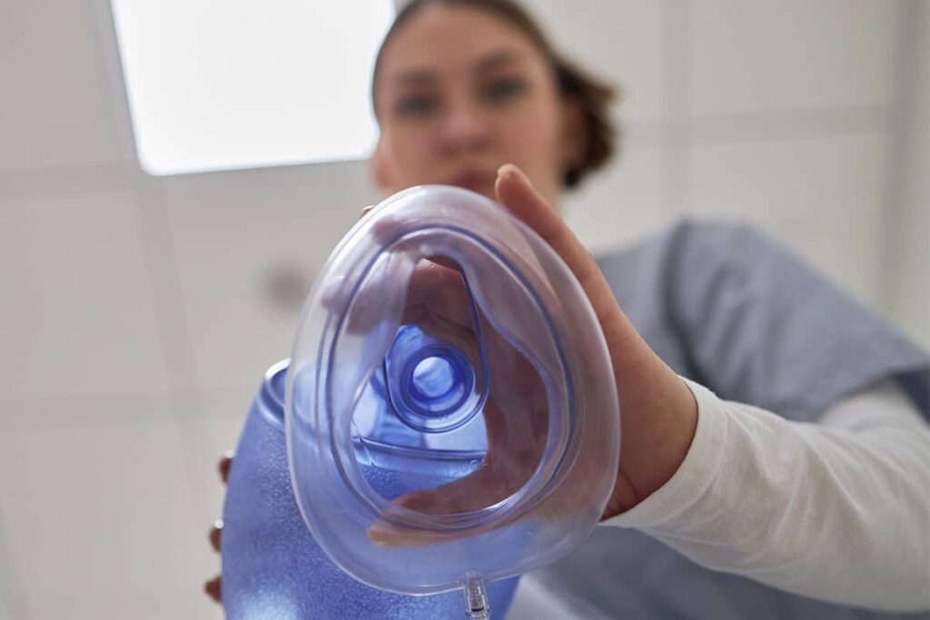 A low-angle view looking up at a medical practitioner in blue scrubs holding a clear plastic anesthesia breathing mask directly over the camera.